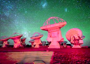 Some of the ALMA antennae bathed in red light. In the background there is the southern Milky Way on the left and the Magellanic Clouds at the top